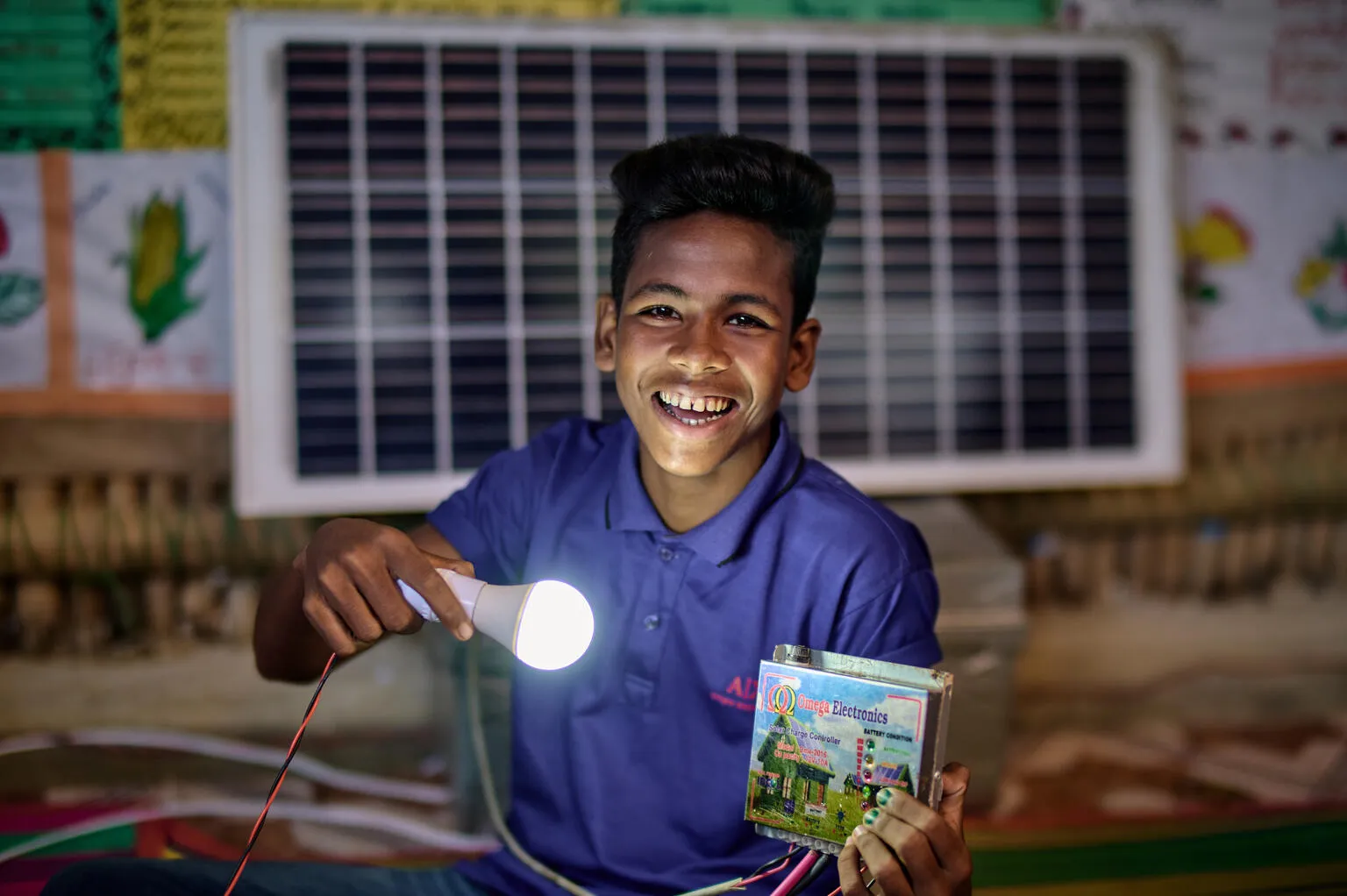 Nur shines a light on a book while smiling and wearing a purple polo shirt at a multi-purpose centre in the Rohingya refugee camps in Cox's Bazar, Bangladesh.