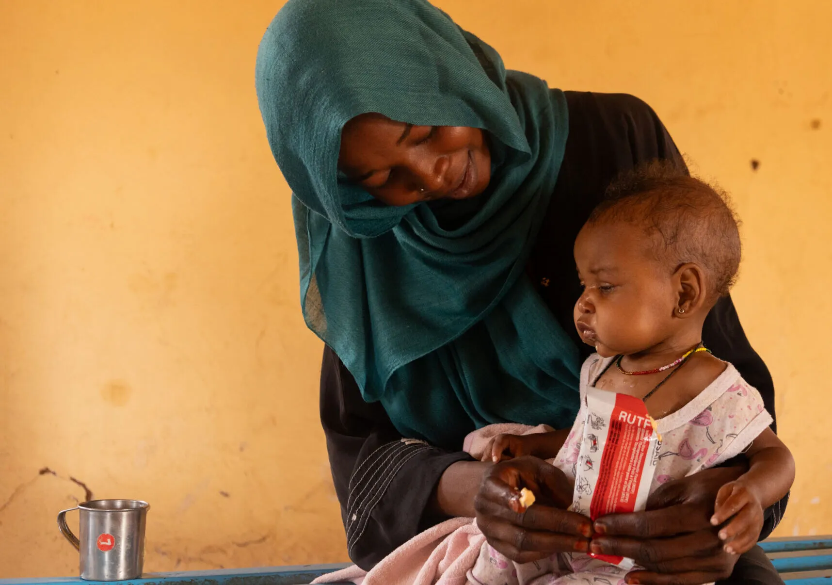 A Sudanese mother looks down lovingly at her infant child sitting on her lap as she feeds them Ready-to-Use Therapeutic Food.